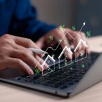 Person arbeitet am Laptop mit eingeblendeten Grafiken von steigenden Häuserpreisen und Prozentzeichen.